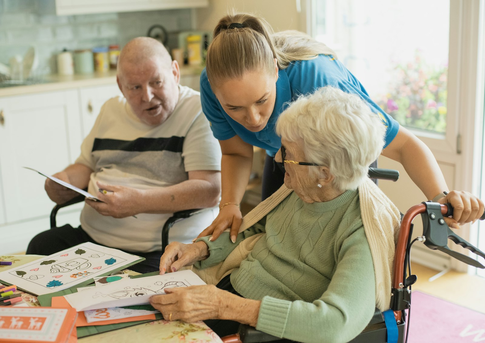 A carer helping two elderly friends with a craft activity at a bright kitchen table